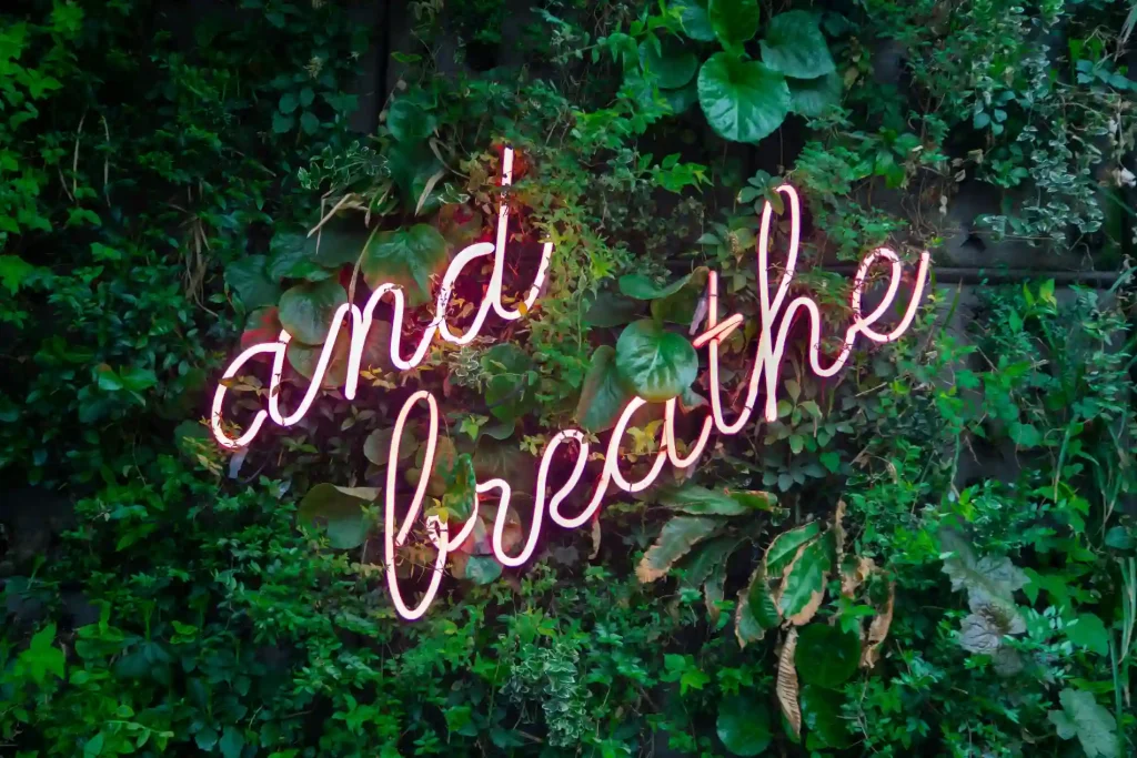 neon sign against green leaves that reads And Breathe