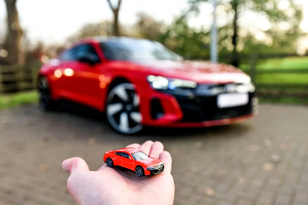 a red Audi toy car on a hand in the foreground in front of the reality of  real dream red audi blurred in the background