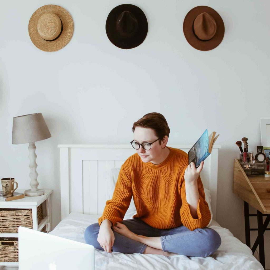 someone sitting on their bed watching something on their laptop, a webinar or zoom call perhaps. there are three hats on the wall and they are holding a book in their hand