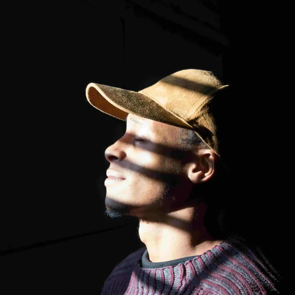 a young man in a baseball cap with sunlight coming through a blind shining and casting shadows on his face. he looks peaceful and mindful