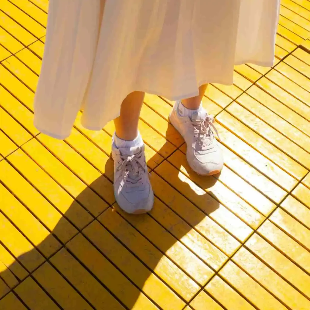 a yellow floor in bright sunshine with someone standing on it wearing a pair of trainers and a white skirt casting a shadow